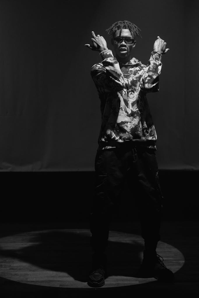 Artistic black and white photo of a performer with dreadlocks making a hand sign on stage.