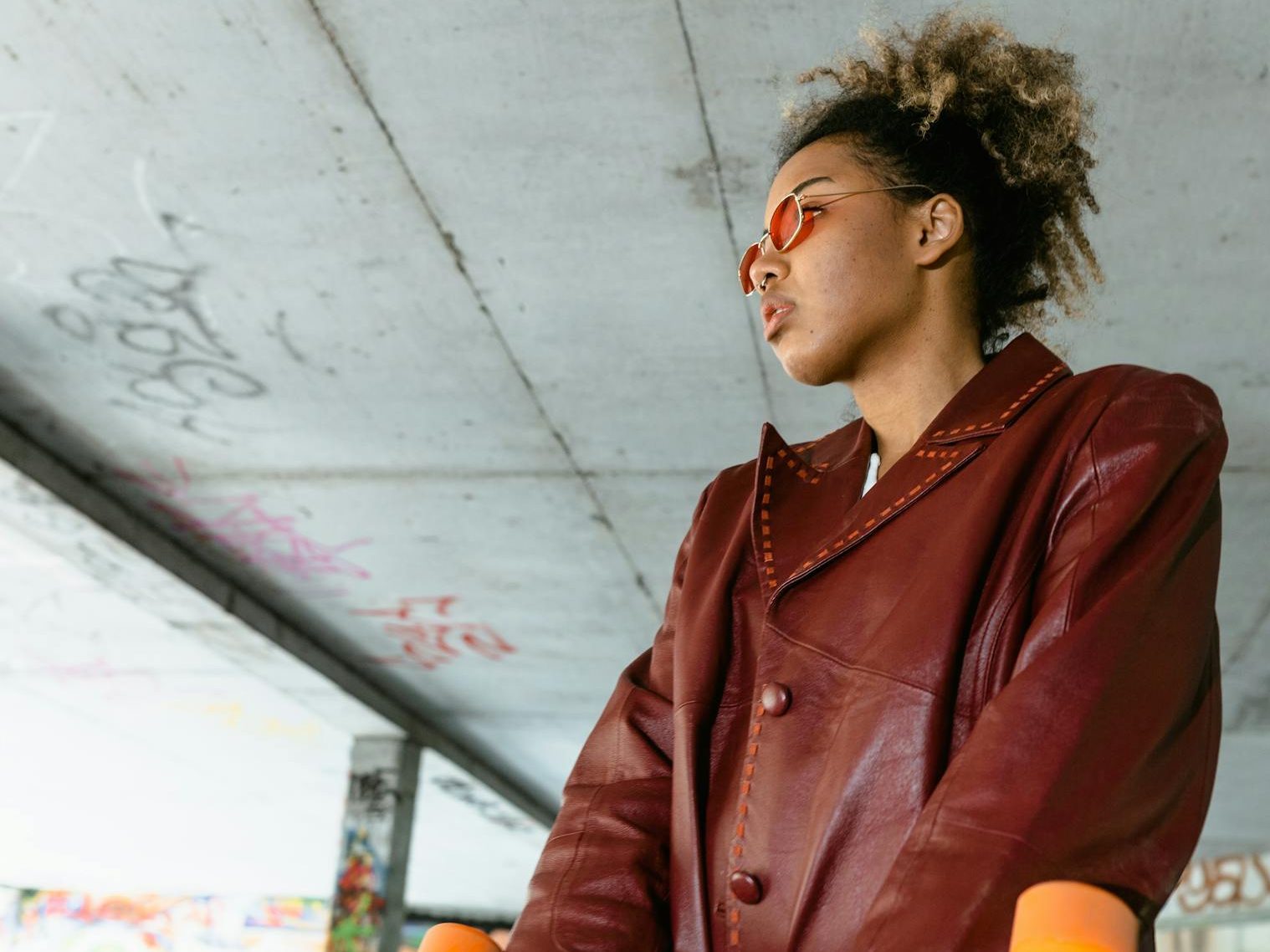 Confident woman in red leather jacket holding a skateboard under a graffiti-filled street overpass.