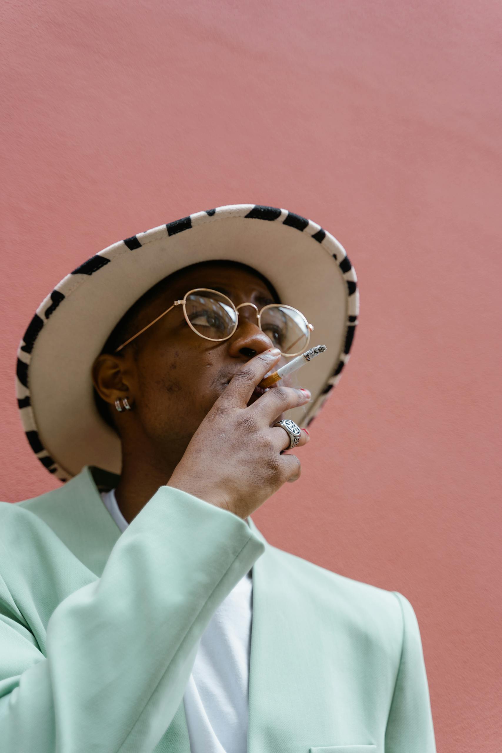 Fashionable man in eyeglasses and hat smoking outdoors.